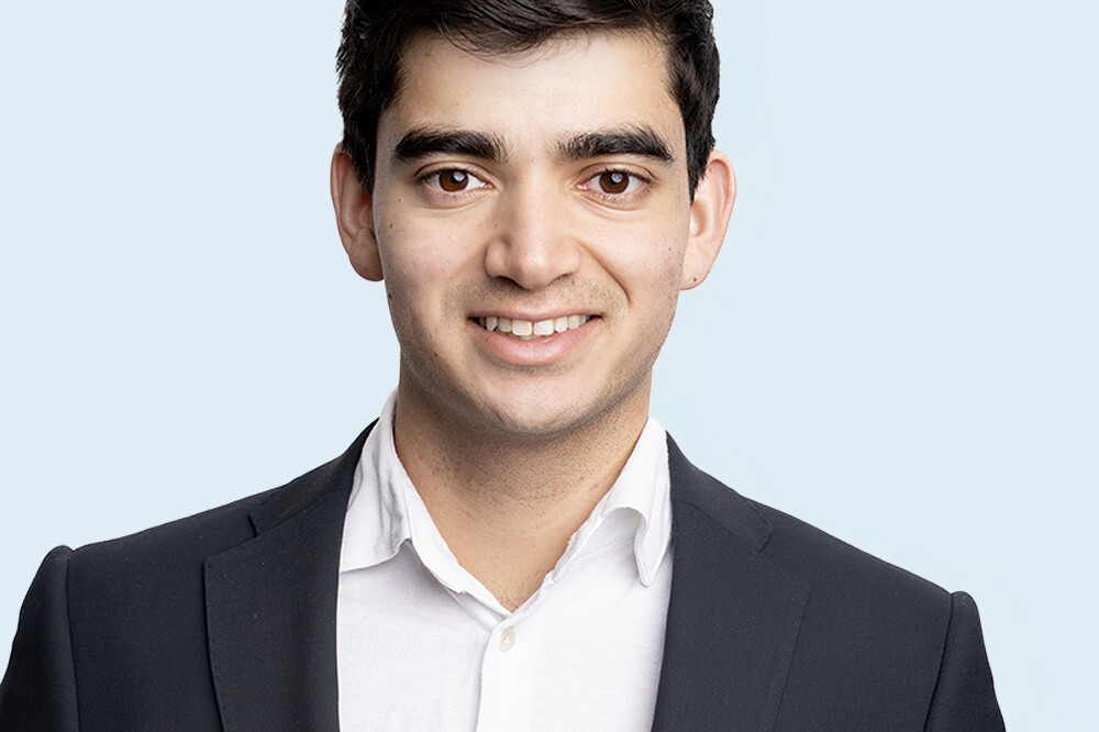 A young man with short dark hair, wearing a dark suit jacket and a white collared shirt, smiles at the camera against a plain light blue background, echoing the polished style of Social Capital Partners' Chief of Staff Joanna Rodrigues.