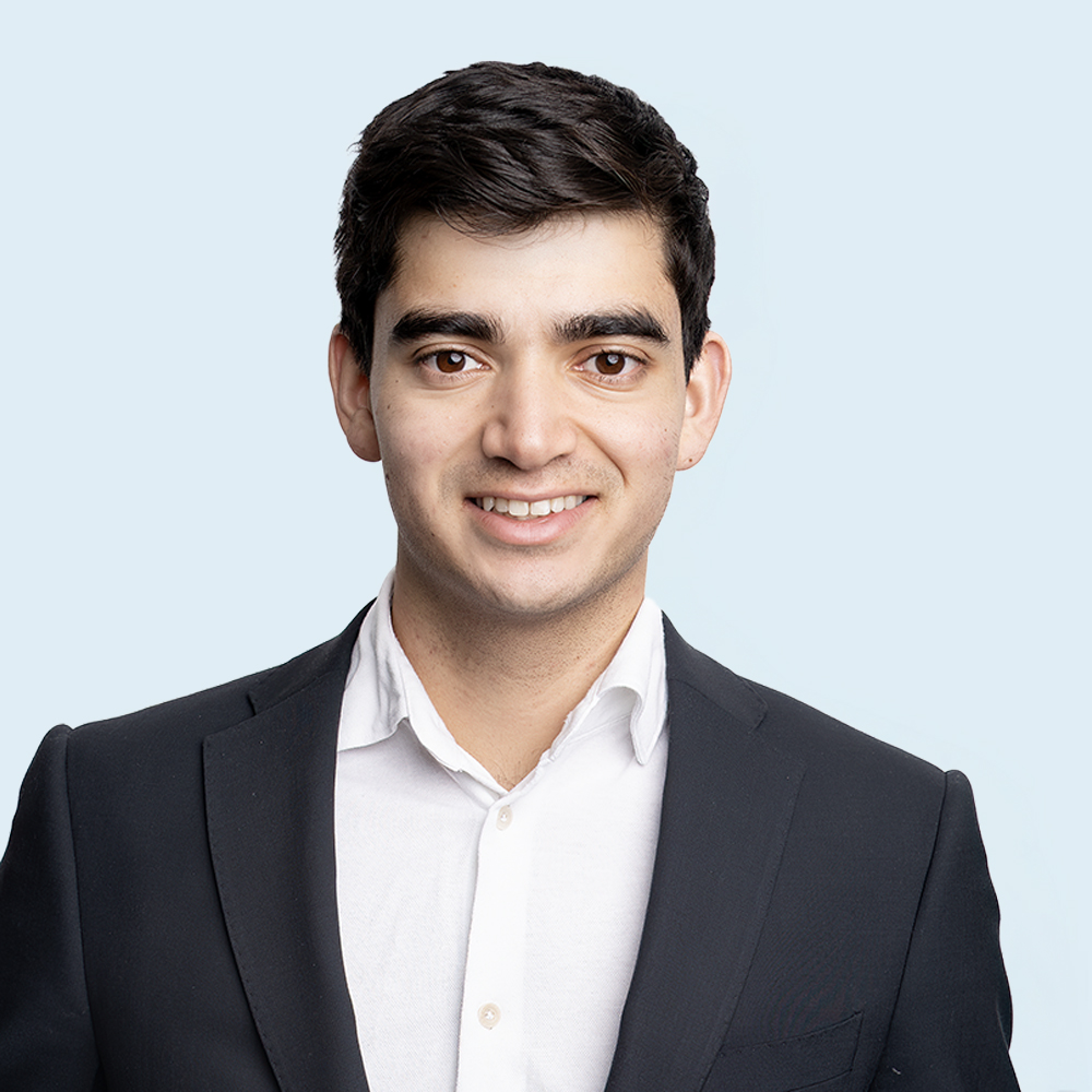 A young man with short dark hair, wearing a dark suit jacket and a white collared shirt, smiles at the camera against a plain light blue background, echoing the polished style of Social Capital Partners' Chief of Staff Joanna Rodrigues.