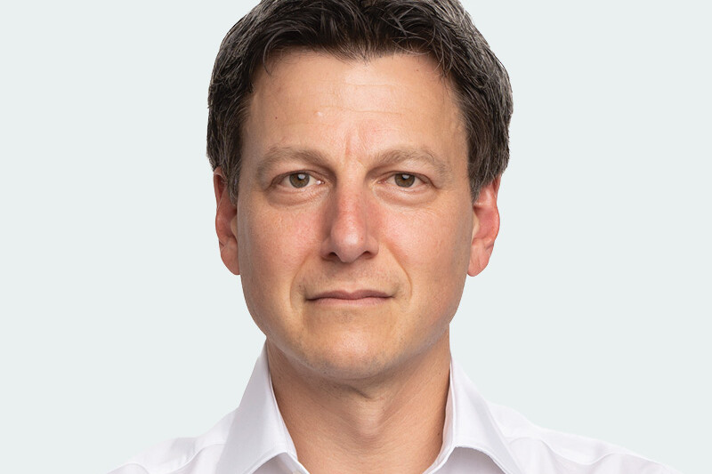 Social Capital Partners' Chair Jon Shell, a man with short dark hair wearing a white collared shirt, looks directly at the camera with a neutral expression against a light gray background.