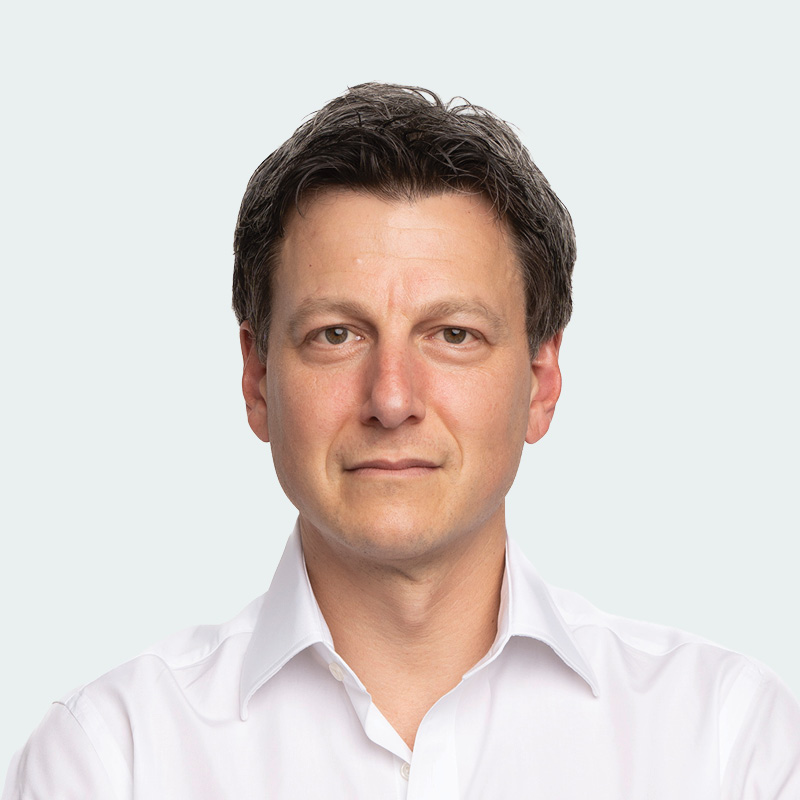 Social Capital Partners' Chair Jon Shell, a man with short dark hair wearing a white collared shirt, looks directly at the camera with a neutral expression against a light gray background.