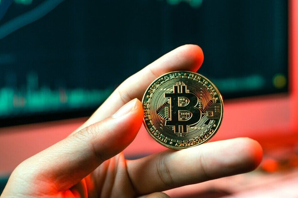 Hand holding a Bitcoin in front of a computer showing digital graphs