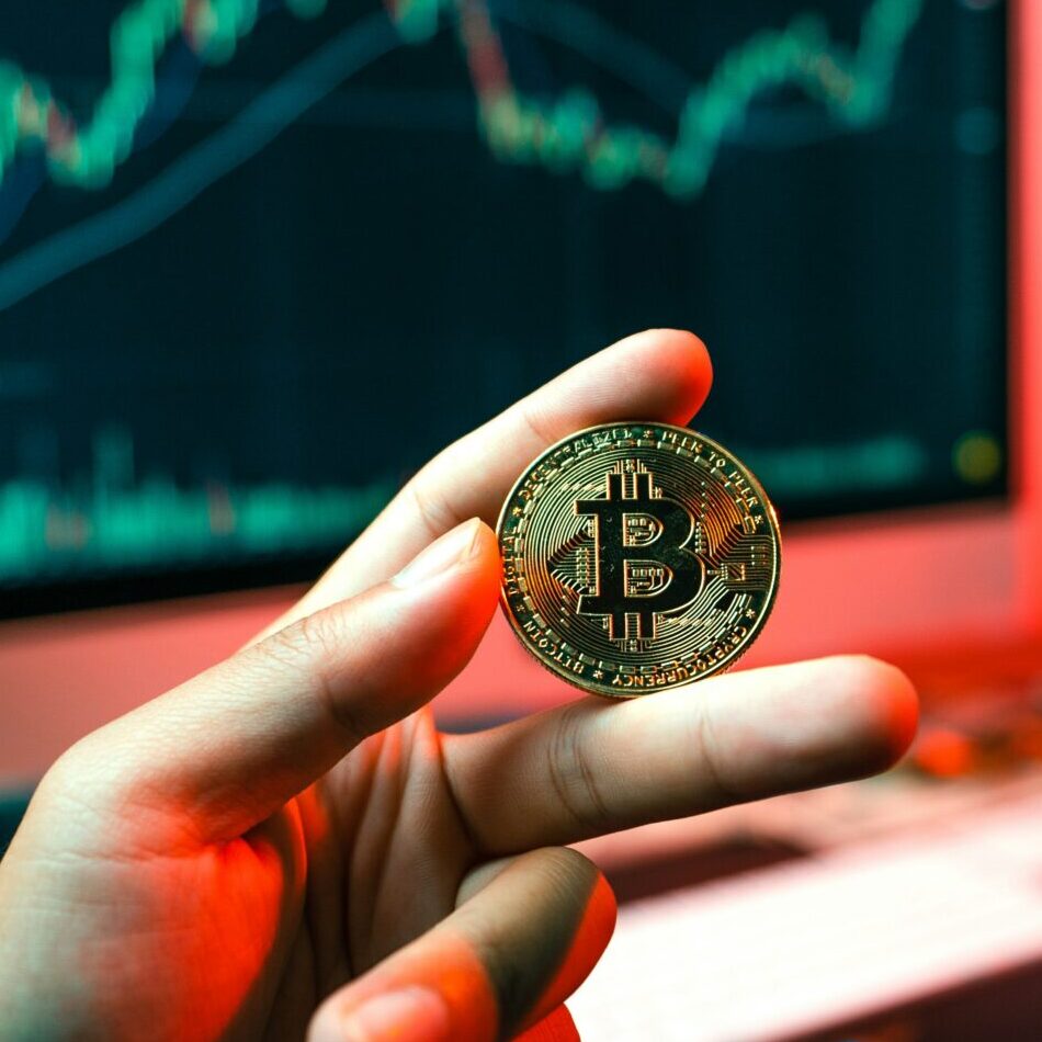 Hand holding a Bitcoin in front of a computer showing digital graphs