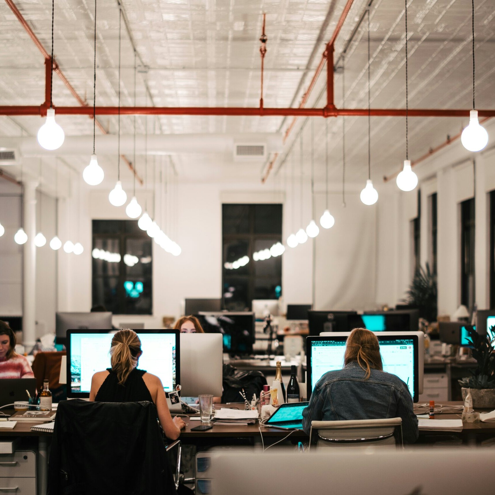 A modern open-plan office in London, Ontario, with several people working at computers. Hanging light bulbs illuminate the space, while large windows brighten the busy workspace—perfect for teams discussing a Rate Drop Rebate opportunity.