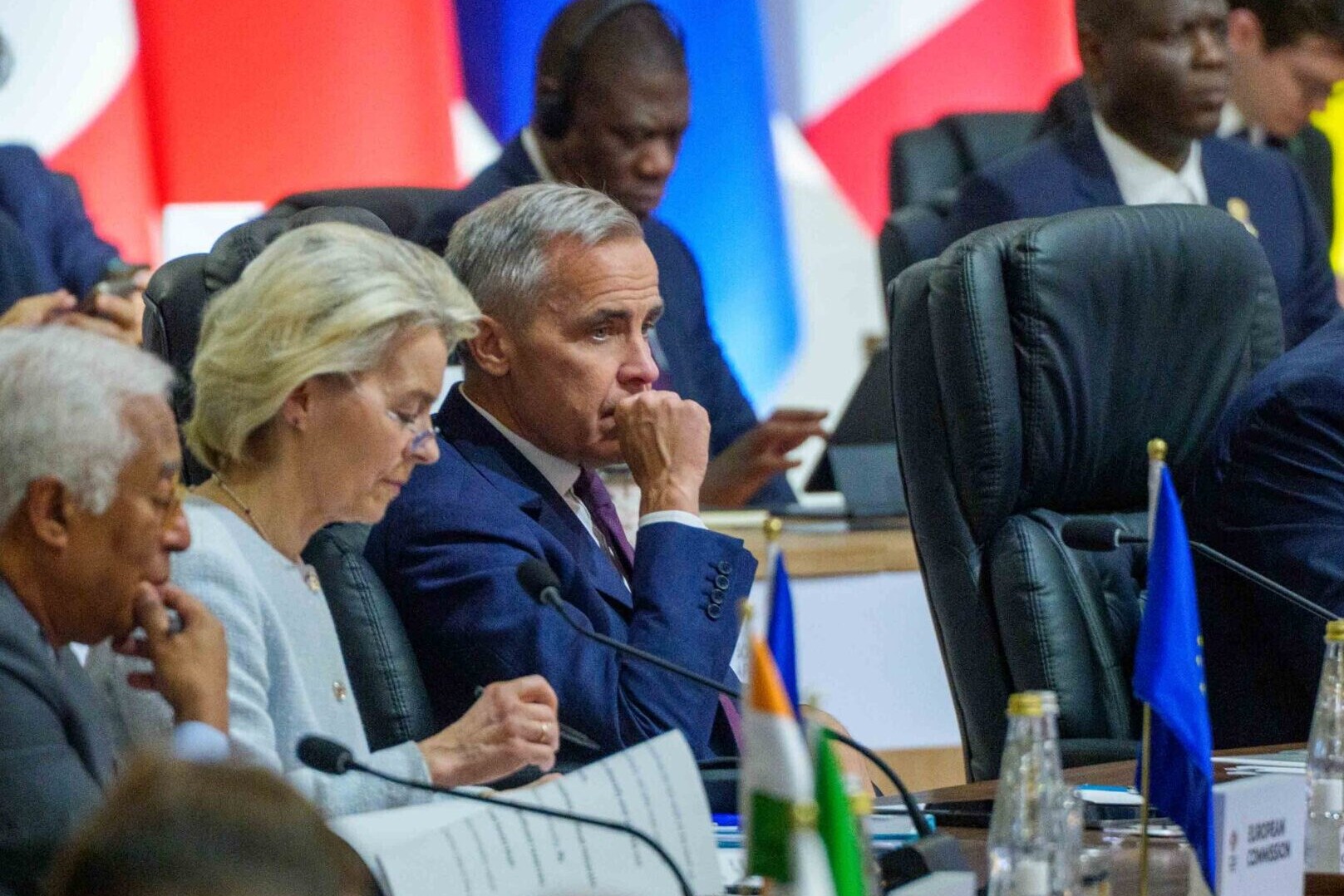 Leaders and delegates sit at a long table during an international summit, with country flags in the background. They appear focused and engaged in discussion, possibly addressing topics like employee ownership trusts FAQs, with documents and nameplates in front of them.