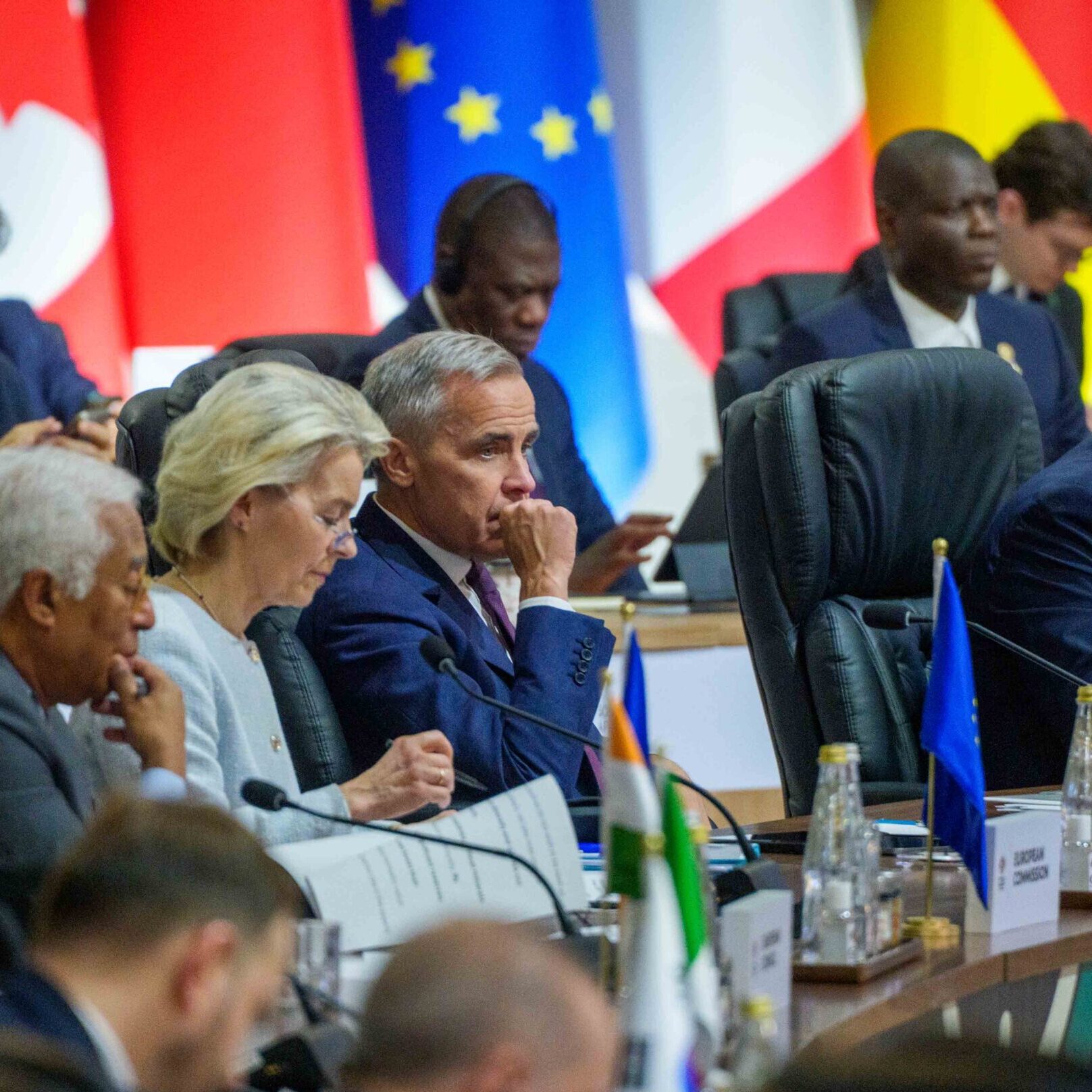 Leaders and delegates sit at a long table during an international summit, with country flags in the background. They appear focused and engaged in discussion, possibly addressing topics like employee ownership trusts FAQs, with documents and nameplates in front of them.