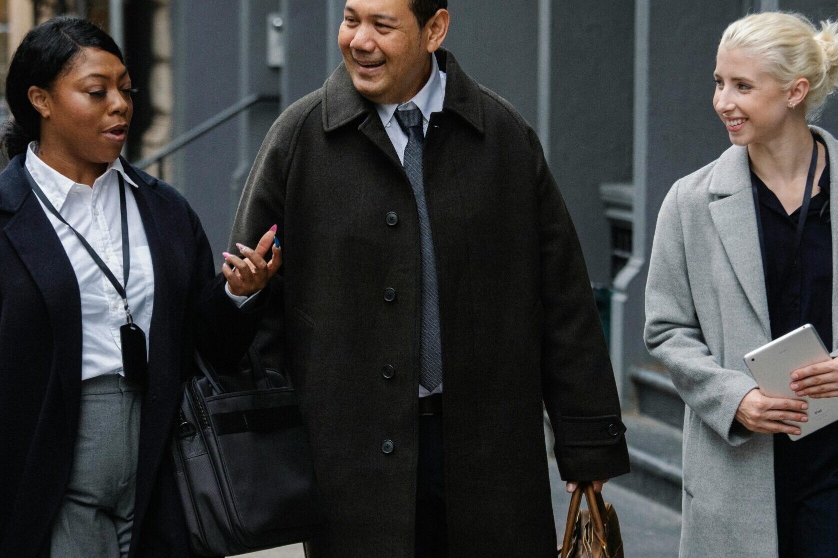 Three professionally dressed people walk together on a city sidewalk, smiling and talking about employee ownership trusts FAQs. One woman holds a notebook, another gestures while speaking, and the man carries a bag.