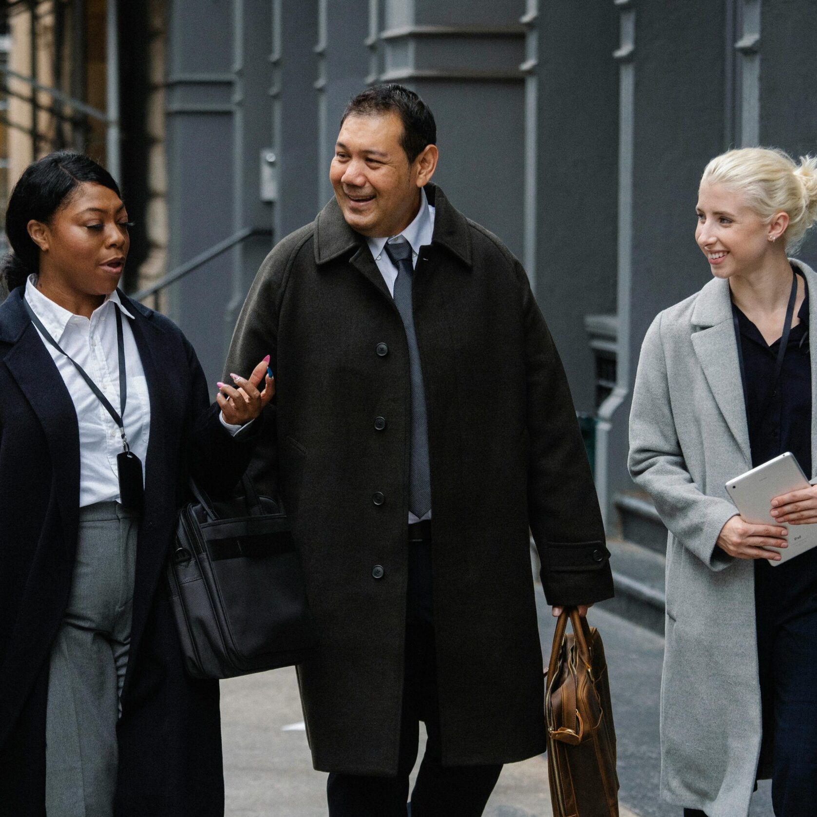 Three professionally dressed people walk together on a city sidewalk, smiling and talking about employee ownership trusts FAQs. One woman holds a notebook, another gestures while speaking, and the man carries a bag.